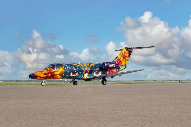 Side view of a Citation CJ3+ business jet on a runway, painted in a multicoloured livery with large flowers and stylised eyes against a blue sky.