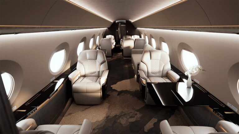 Forward cabin of a Gulfstream G700 business jet featuring cream leather seats, dark wood cabinetry, gold accents, oval windows and a high-gloss black table.