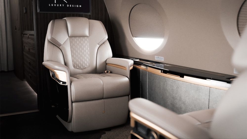 Close-up of a champagne-taupe leather aircraft seat with diamond-quilted inserts and dark wood side bulkhead, positioned beside a cabin window.