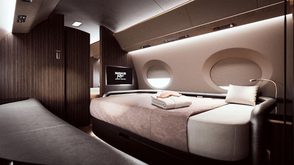 Private aircraft bedroom suite with a made-up bed in neutral tones, dark wood bulkheads with embedded screen displaying the Natalie Rodríguez Luxury Design logo.