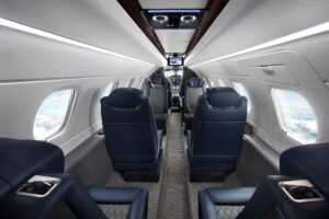 The interior cabin of the Phenom 300EV, showing navy leather seats with diamond stitching, large windows and a central aisle looking towards the cockpit