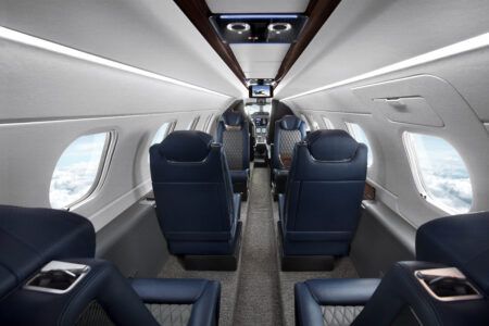The interior cabin of the Phenom 300EV, showing navy leather seats with diamond stitching, large windows and a central aisle looking towards the cockpit