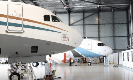 Two Airbus Corporate Jet aircraft parked nose-first inside a maintenance hangar, one in the foreground on jacks.
