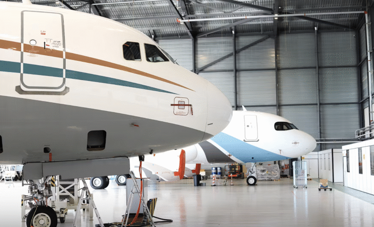 Two Airbus Corporate Jet aircraft parked nose-first inside a maintenance hangar, one in the foreground on jacks.