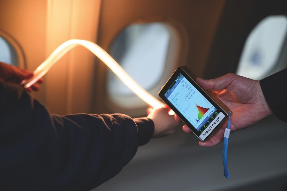 Hands holding an illuminated LED light strip next to a handheld spectrometer displaying light spectrum data inside a business jet cabin
