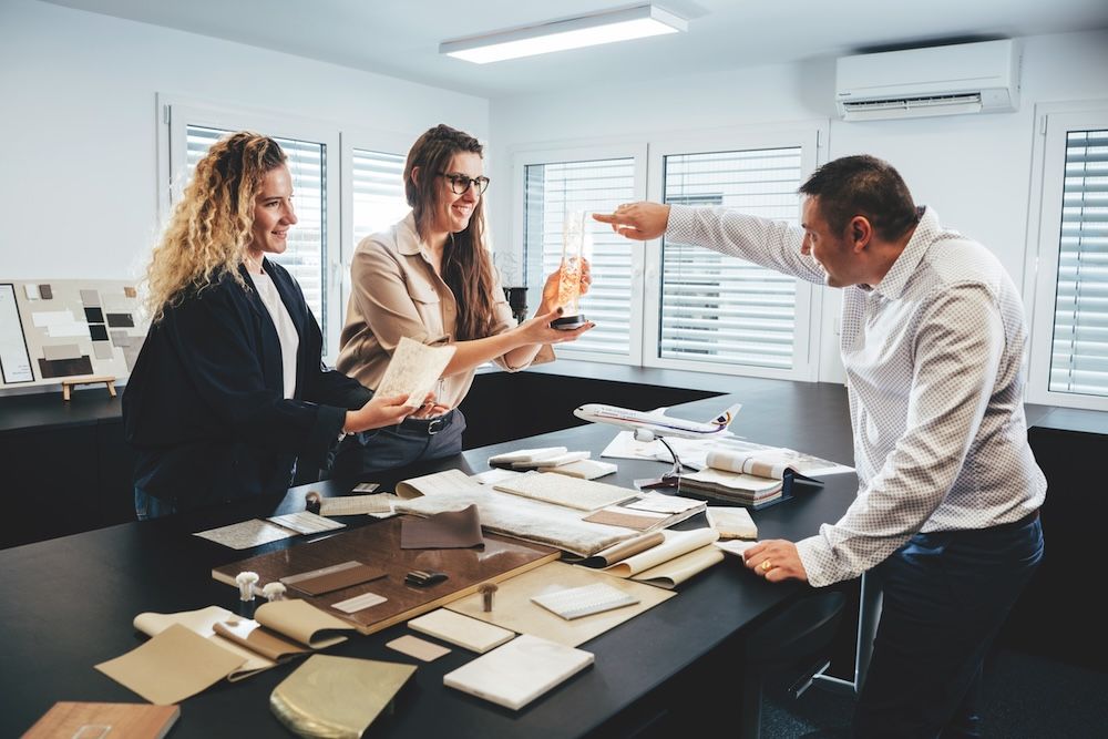 Three AMAC Aerospace design team members reviewing carpet, leather and veneer samples at a desk, with a model aircraft and material swatches spread across the table.