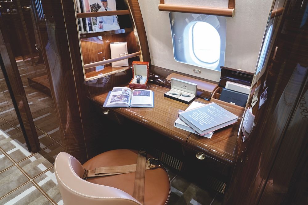 Wood-veneer cabin desk and cream leather seat in a VIP business jet interior, with amenities laid out on the desk.
