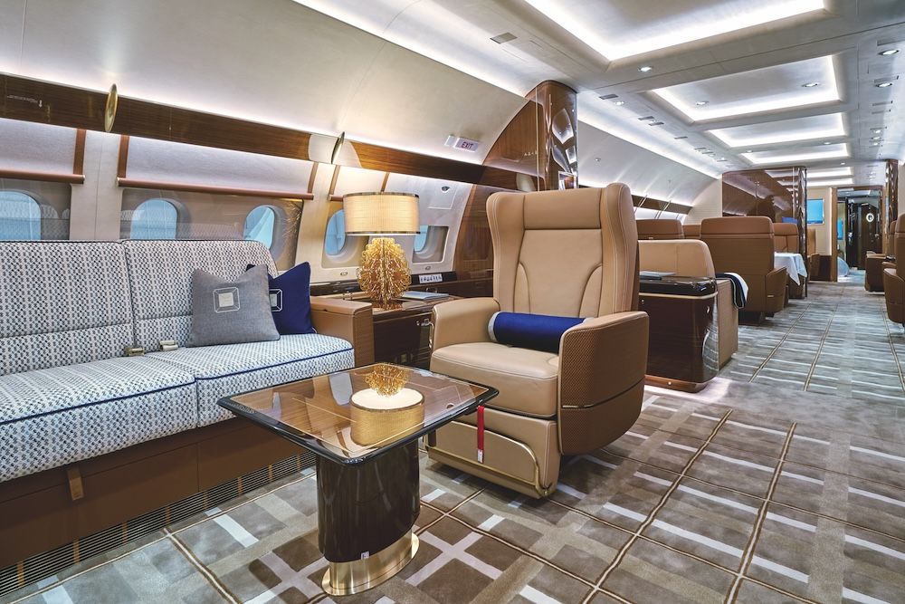 Business jet cabin interior with tan leather reclining seats, a patterned divan, gloss veneer coffee table, and gold table lamp