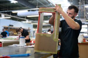A technician in a JCB Aero workshop holds up a red-edged composite cabin monument panel, inspecting the honeycomb core and bonded surface finish.