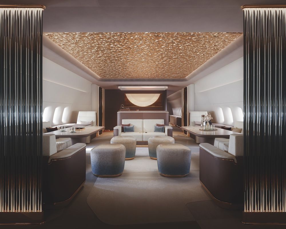 VIP business jet cabin lounge with a textured gold ceiling panel, cream upholstered seating, ottomans and a bar area, whole scene framed by fluted columns.