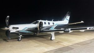 A white TBM 980 single-engine turboprop with blue and black livery parked on an apron at night