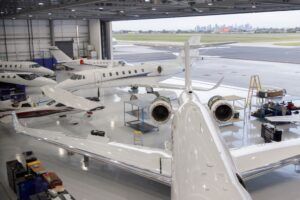 Wide interior view of an aircraft maintenance hangar with several business jets, hangar doors open to a runway beyond