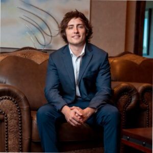 Brody Speno, wearing a navy suit and white open-necked shirt, sits on a brown leather studded sofa with an abstract artwork on the wall behind