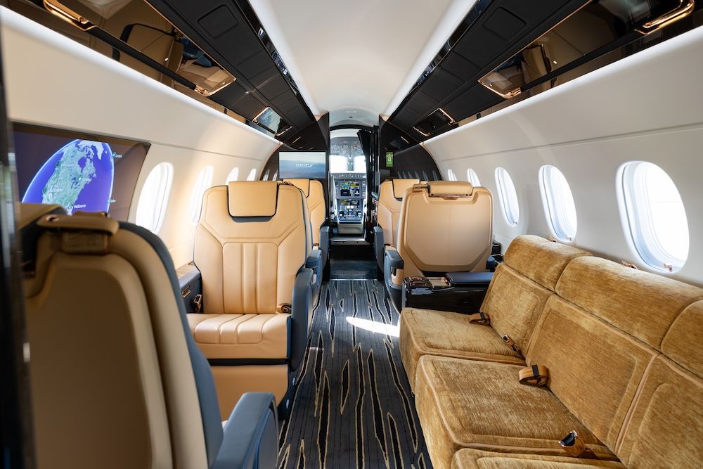 Forward view of the Praetor 600E cabin looking forward, showing tan leather seats, and a side-facing divan facing a large sidewall display screen