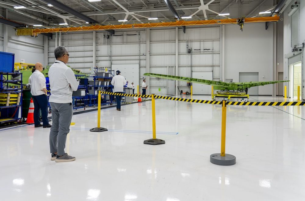 Employees observing a wing structure being transported on a cart inside a manufacturing facility