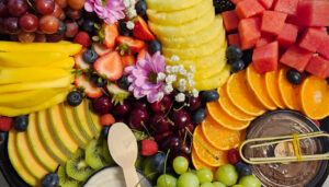 Close-up of a fruit platter with mango, strawberries, pineapple, watermelon, orange slices, cherries, cantaloupe, kiwi, grapes, cherries, raspberries and blueberries, decorated with pink and white flowers, next to a Nutella dip