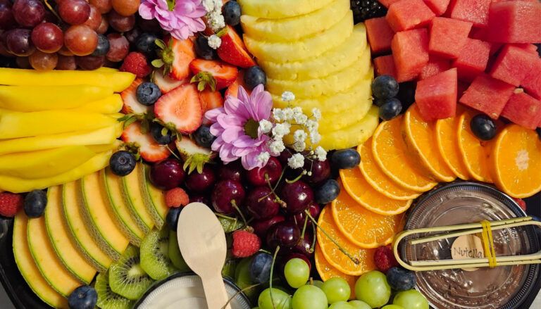 Close-up of a fruit platter with mango, strawberries, pineapple, watermelon, orange slices, cherries, cantaloupe, kiwi, grapes, cherries, raspberries and blueberries, decorated with pink and white flowers, next to a Nutella dip