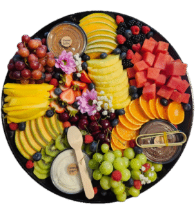 Overhead view of a large round black tray of sliced fresh fruit and berries with labelled pots of almond butter, caramel yogurt and Nutella, plus a wooden spoon and bamboo tongs