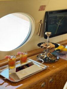 Business jet cabin credenza with a silver tray holding two iced drinks garnished with citrus and a napkin-wrapped bowl of berries, beside a tiered stand of biscuits, nuts and dried orange, under an oval window and a moving-map screen