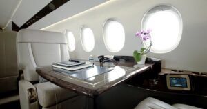 Business jet cabin with cream leather seats, a polished wood table, oval windows and a touchscreen control panel built into the sideledge