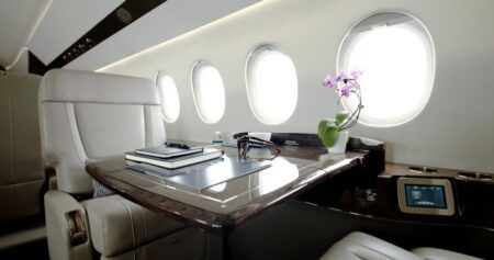 Business jet cabin with cream leather seats, a polished wood table, oval windows and a touchscreen control panel built into the sideledge