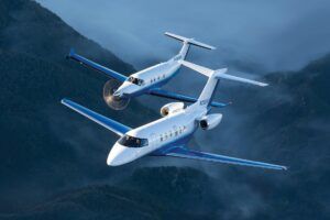 A white-and-blue Pilatus PC-24 business jet and a Pilatus PC-12 single-engine turboprop fly in close formation above forested mountains