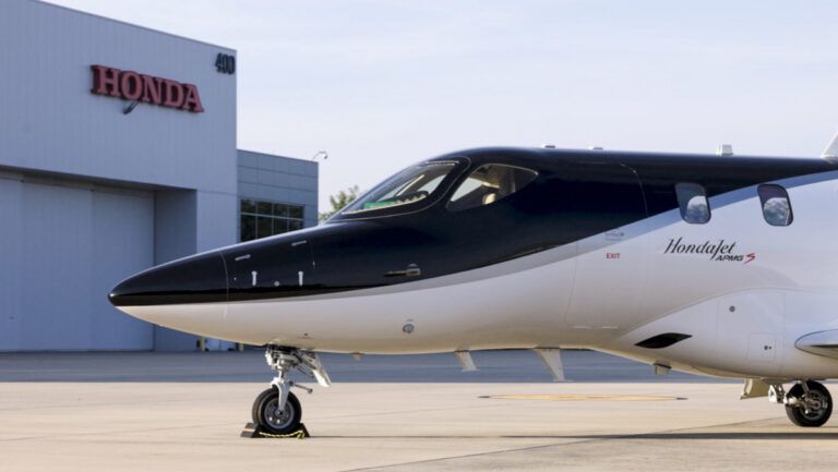 The nose and forward fuselage of a HondaJet, painted with a glossy black upper section and white lower section in front of a grey hangar with a red Honda sign
