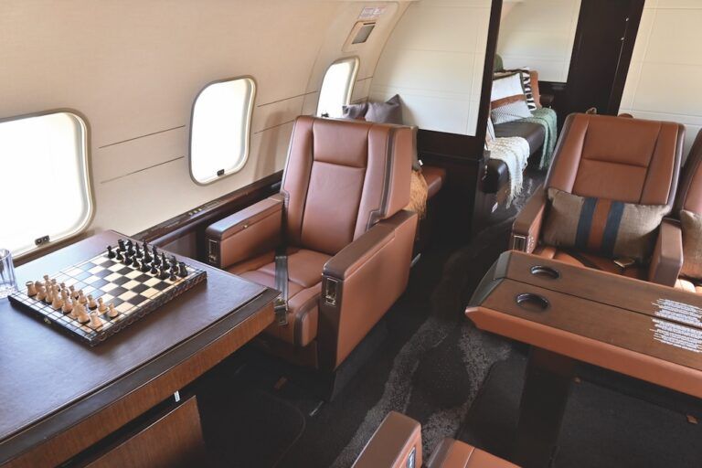 A business jet cabin with tan leather club seats, veneered sideledges and tables, and a chess set laid out on a table by the windows. Behind them, a divan in the aft section is dressed with patterned cushions and throws.