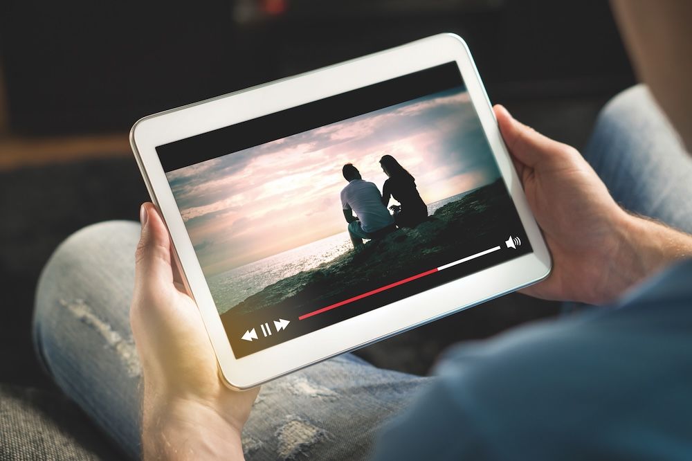 Hands holding a tablet that is streaming a video of a couple sitting on a rock by the sea