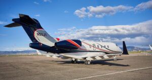A business jet with a white, navy and red livery parked on an apron, with mountains and blue sky behind