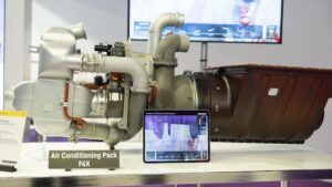 A silver air-conditioning pack labelled "Air Conditioning Pack F6X" on a white display table beside a tablet showing a digital model of the unit, with a screen in the background.