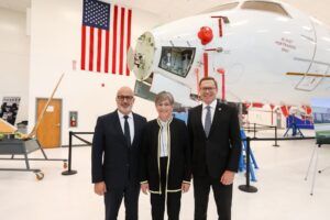 Two men in dark suits and a woman in a black jacket with gold trim standing in front of a white aircraft with an open nose section, with a large US flag on the hangar wall behind