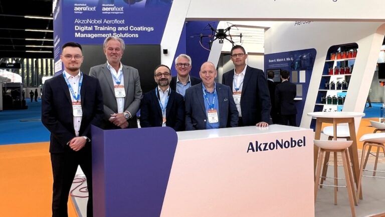 Aerospace launch for AkzoNobel’s Mixit digital colour-retrieval system Six men in business attire wearing lanyards stand behind a white and purple AkzoNobel Aerospace Coatings stand at a trade show.