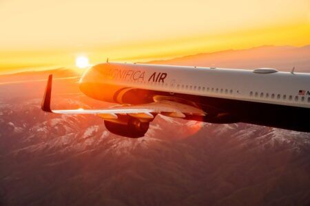 A white aircraft in Magnifica Air livery flying over snow-dusted mountains at sunset, with the sun low on the horizon