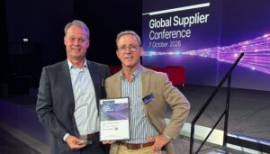Two men on a stage in front of a screen reading "Global Supplier Conference 7 October 2026", one holding a glass trophy and the other a framed Game Changer Award certificate.