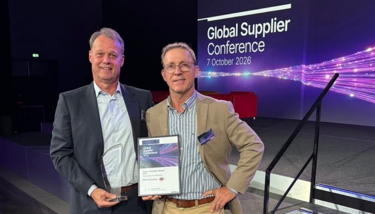 Two men on a stage in front of a screen reading "Global Supplier Conference 7 October 2026", one holding a glass trophy and the other a framed Game Changer Award certificate.