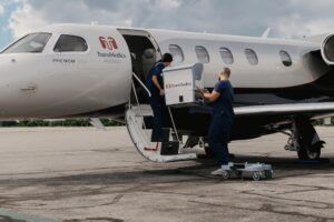 Two medical staff in navy scrubs load a TransMedics organ transport unit into a TransMedics Aviation Embraer Phenom 300E business jet through its open airstair door on the apron